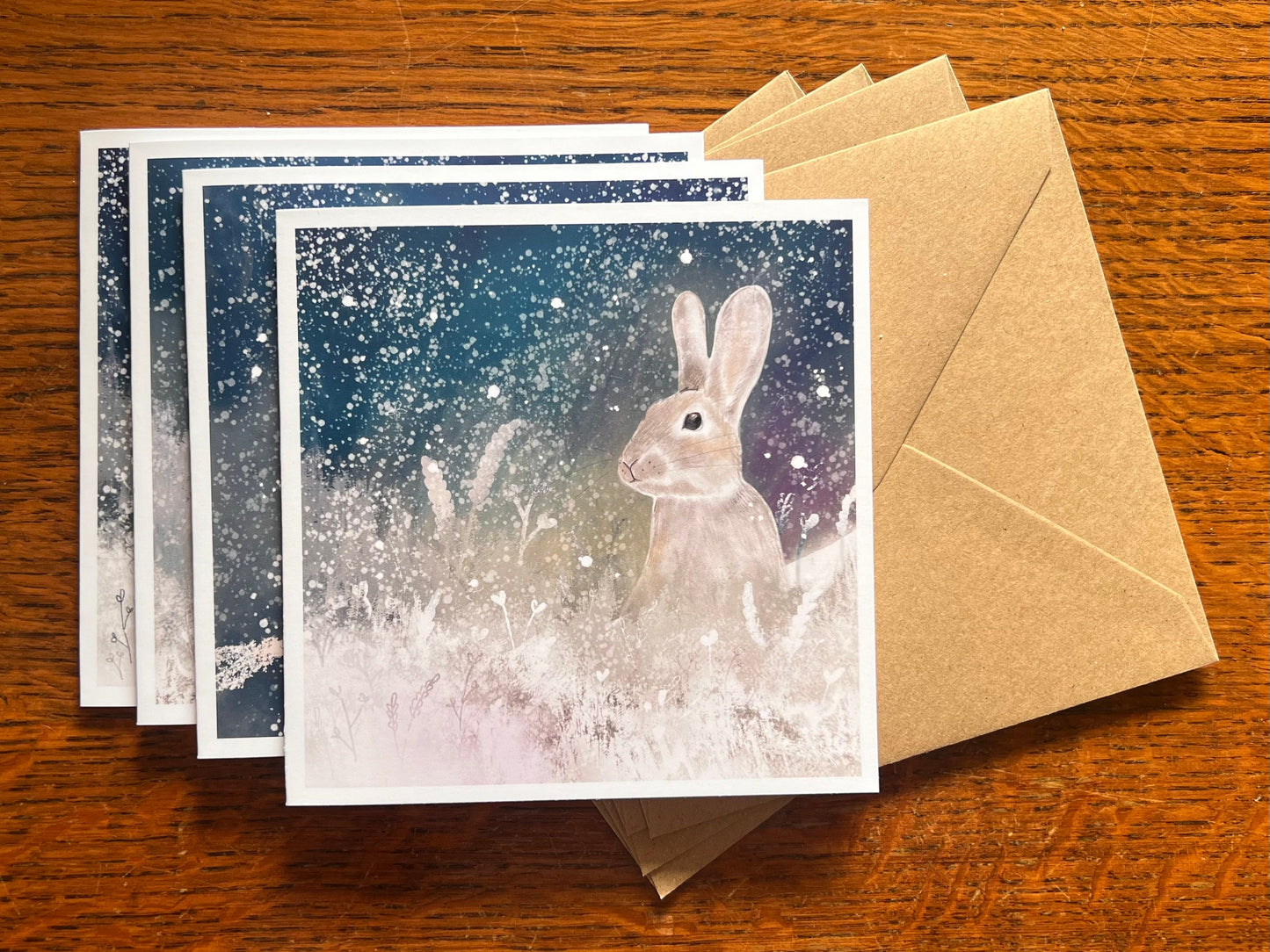Set of four winter animal greeting cards stacked on top of recycled kraft envelopes on a wooden tabletop, with the top card featuring an illustrated silvery grey rabbit with raised ears sitting among snowy foliage under a magical blue, green and purple winter night sky with falling snow, with fox, owl and stag cards visible beneath.