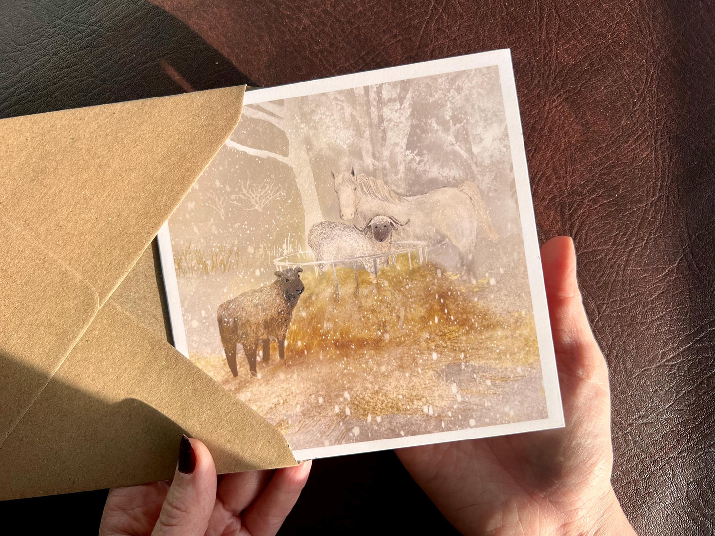 Premium Christmas card featuring a magical winter farm scene with a white horse and two sheep gathered around a hay bale in the snow. The soft taupe pink background represents a snowy evening sunset. The card has no text and is shown in close-up being held in two hands. Ideal Christmas card for animal lovers, horse enthusiasts and farm scene greeting card collectors. Printed on thick 312gsm fine art card by a UK independent artist.