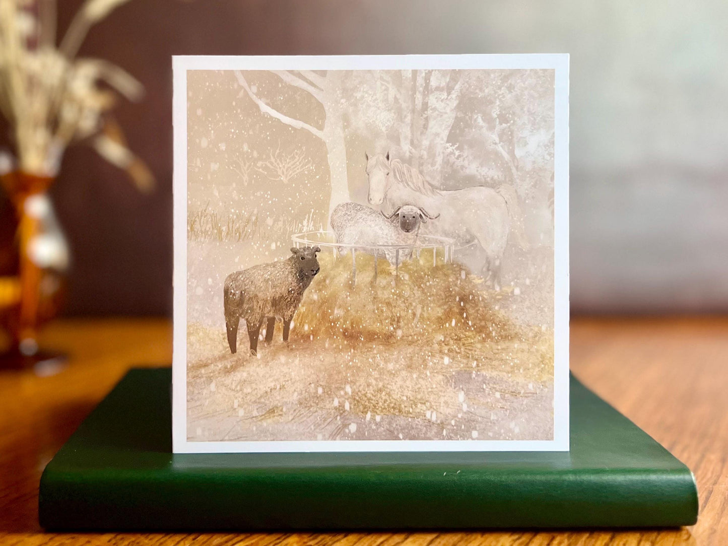Premium Christmas card featuring a magical winter farm scene with a white horse and two sheep standing in the snow around a hay bale. The soft taupe pink sunset background creates a peaceful festive feel. The card is shown standing on a green leatherette book in a styled display. Ideal Christmas card for horse lovers, animal enthusiasts and farm scene greeting card collectors. Printed on thick 312gsm fine art card by a UK independent artist.