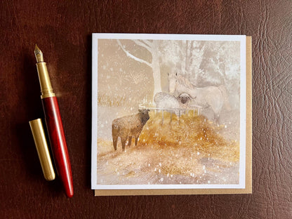 Premium Christmas card featuring a magical winter farm scene with a white horse and two sheep standing around a hay bale in the snow. The taupe pink sunset background creates a soft festive atmosphere. The card is shown laid flat with a recycled kraft envelope and fountain pen on a brown leatherette tabletop. Ideal Christmas card for horse lovers, animal enthusiasts and farm scene greeting card collectors. Printed on thick 312gsm fine art card by a UK independent artist.