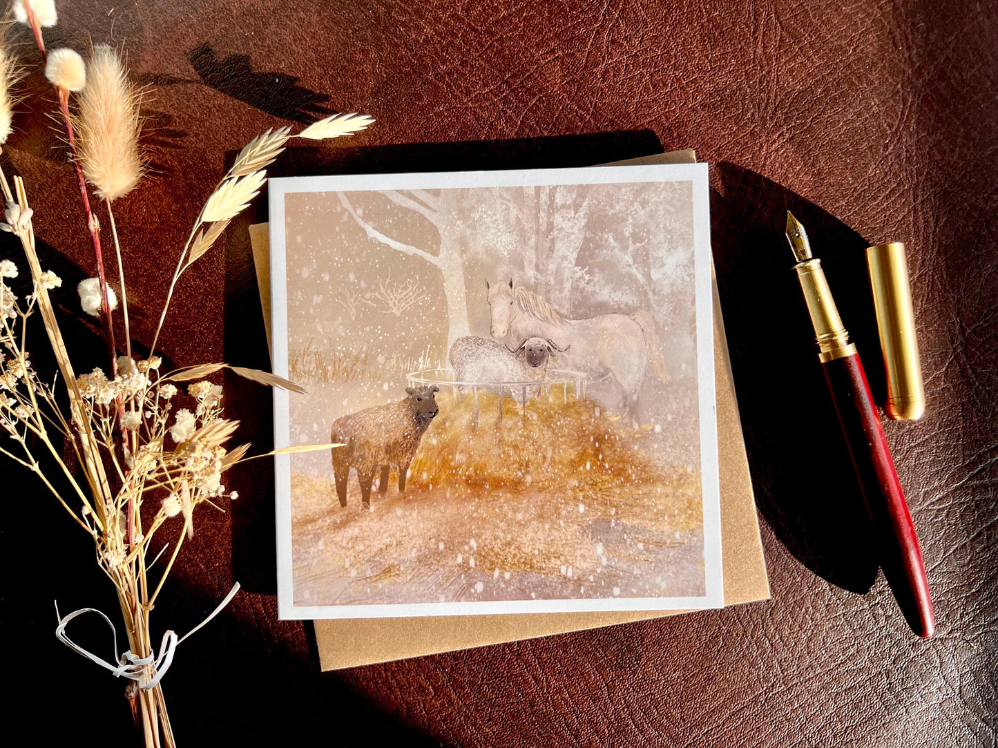 Premium Christmas card featuring a magical winter farm scene with a white horse and two sheep standing around a hay bale in the snow. The soft taupe pink sunset background creates a gentle festive feel. The card is shown laid flat with a recycled kraft envelope, wooden fountain pen and dried flowers on a leatherette tabletop, with sunlight casting soft shadows. Ideal Christmas card for horse lovers, animal enthusiasts and farm scene greeting card collectors. Printed on thick 312gsm fine art card