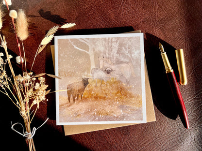 Premium Christmas card featuring a magical winter farm scene with a white horse and two sheep standing around a hay bale in the snow. The soft taupe pink sunset background creates a gentle festive feel. The card is shown laid flat with a recycled kraft envelope, wooden fountain pen and dried flowers on a leatherette tabletop, with sunlight casting soft shadows. Ideal Christmas card for horse lovers, animal enthusiasts and farm scene greeting card collectors. Printed on thick 312gsm fine art card