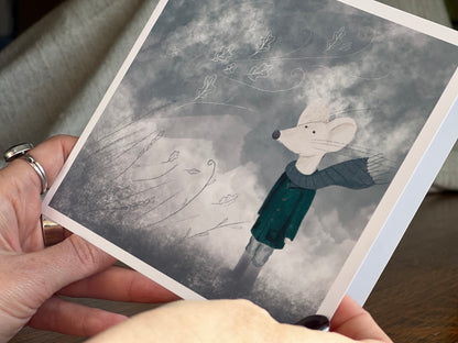 Premium winter greeting card featuring a storybook-style illustration of a white mouse wearing a green scarf and duffle coat in a windy blue-grey winter scene. The card has no text and is shown in close-up being held in two hands, highlighting the front design. Ideal for Christmas, winter greetings or keeping in touch during the colder months. Printed on thick 312gsm fine art card by a UK independent artist.