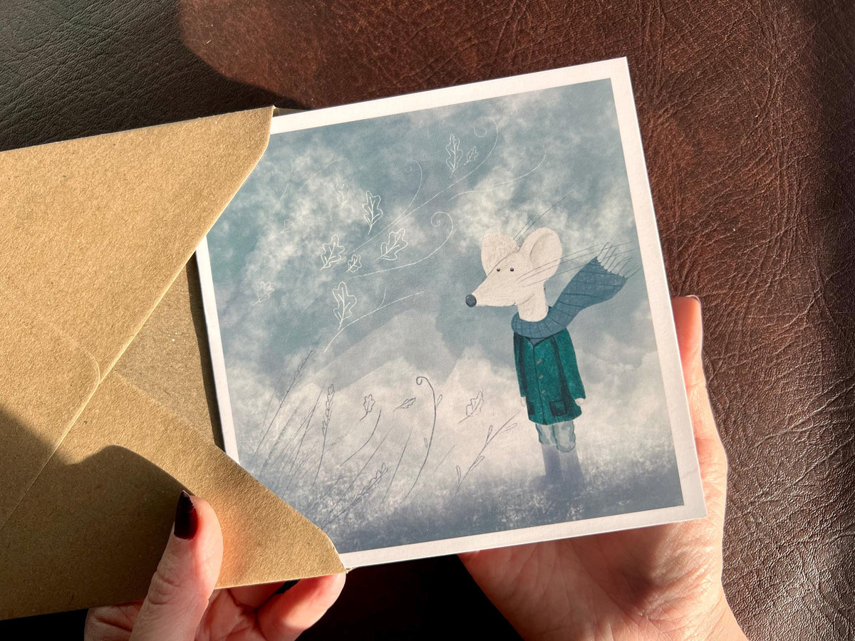 Premium winter greeting card featuring a storybook-style illustration of a white mouse wearing a green scarf and duffle coat, standing in a windy blue-grey winter scene. The card has no text and is shown in close-up being held in two hands. Ideal seasonal greeting card for Christmas, winter birthdays or keeping in touch during the colder months. Printed on thick 312gsm fine art card by a UK independent artist.