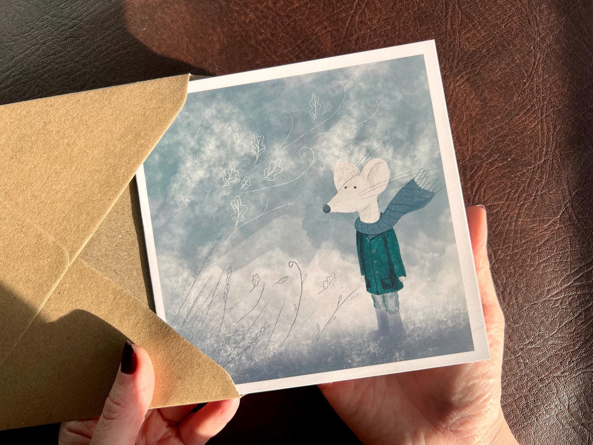 Premium winter greeting card featuring a storybook-style illustration of a white mouse wearing a green scarf and duffle coat, standing in a windy blue-grey winter scene. The card has no text and is shown in close-up being held in two hands. Ideal seasonal greeting card for Christmas, winter birthdays or keeping in touch during the colder months. Printed on thick 312gsm fine art card by a UK independent artist.