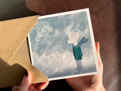 Premium winter greeting card featuring a storybook-style illustration of a white mouse wearing a green scarf and duffle coat, standing in a windy blue-grey winter scene. The card has no text and is shown in close-up being held in two hands. Ideal seasonal greeting card for Christmas, winter birthdays or keeping in touch during the colder months. Printed on thick 312gsm fine art card by a UK independent artist.