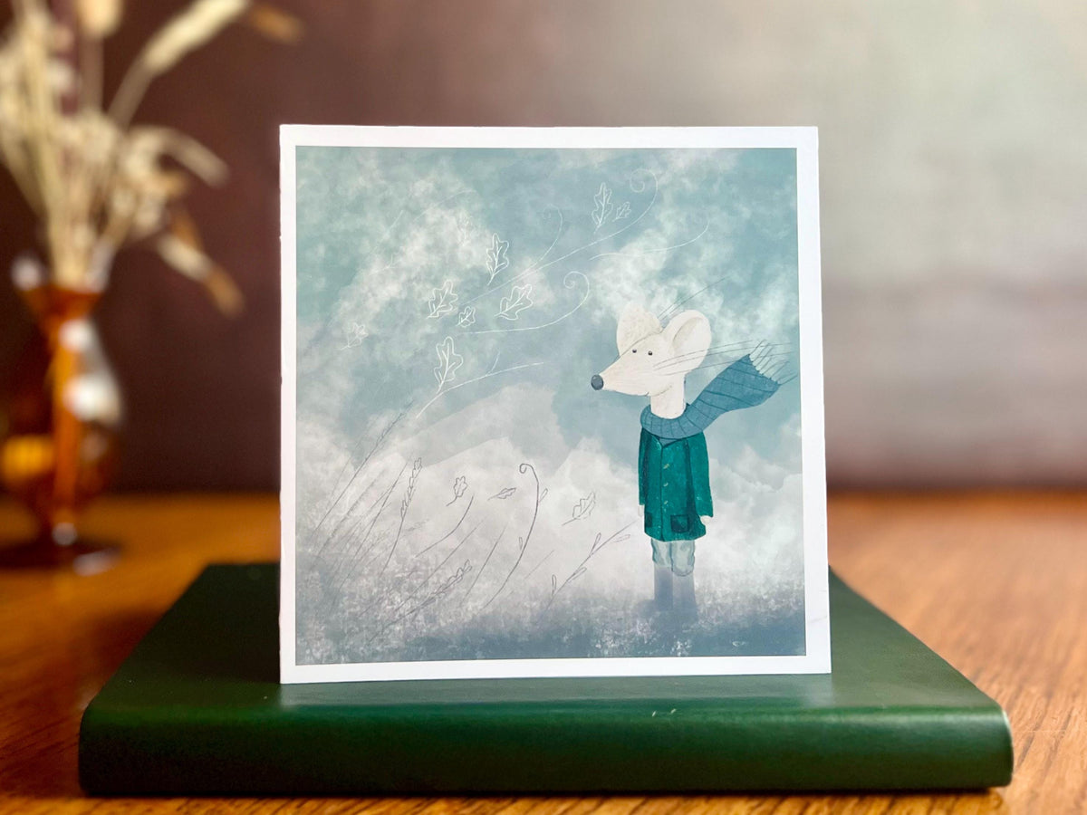 Premium winter greeting card featuring a storybook-style illustration of a white mouse wearing a green scarf and duffle coat in a windy blue-grey winter scene. The card has no text and is shown standing on a book in a styled display. Ideal seasonal greeting card for Christmas, winter greetings or keeping in touch during the colder months. Printed on thick 312gsm fine art card by a UK independent artist.