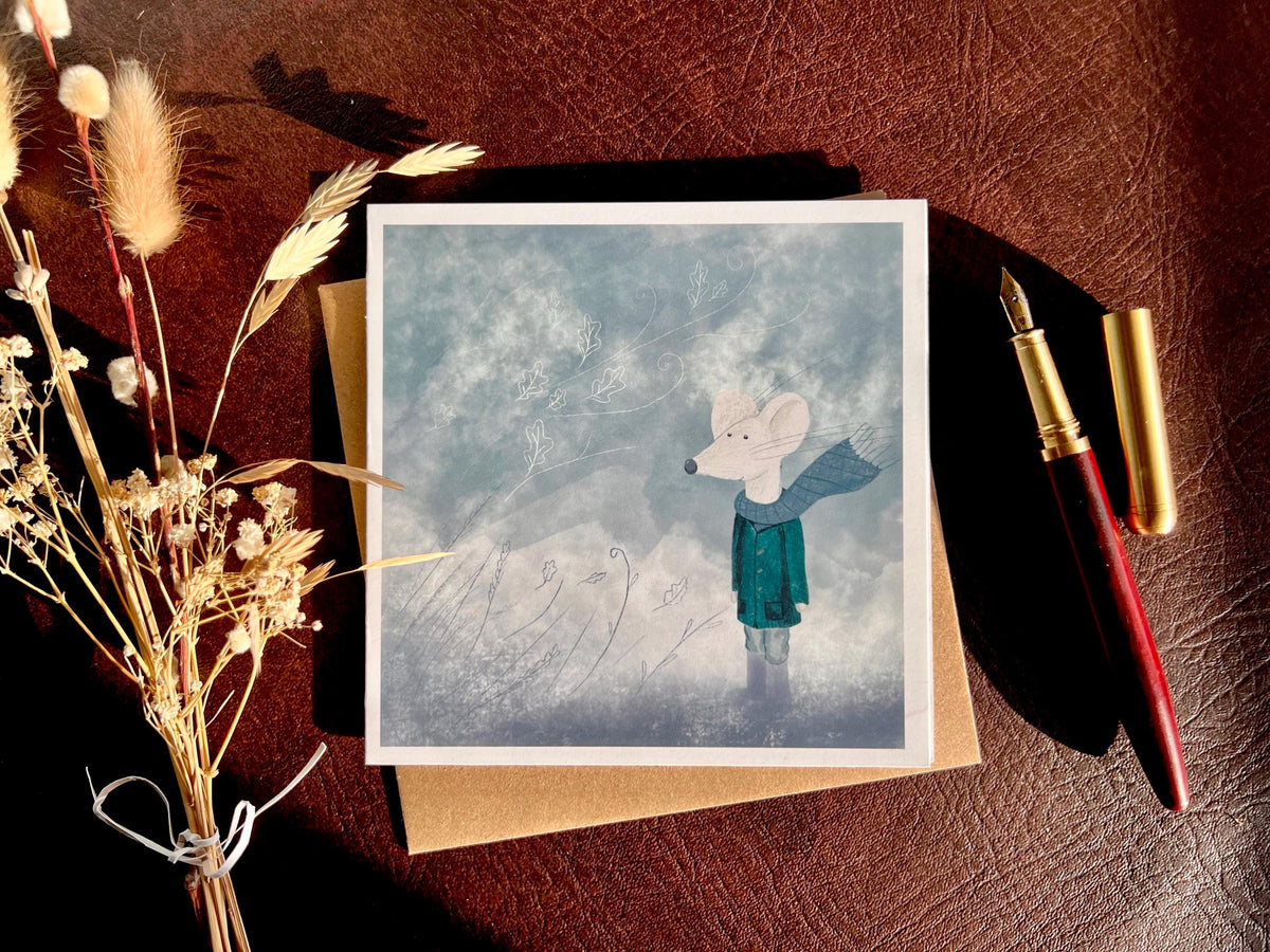 Premium winter greeting card featuring a storybook-style illustration of a white mouse wearing a green scarf and duffle coat in a windy blue-grey winter scene. The card has no text and is shown laid flat on a tabletop. Ideal seasonal greeting card for Christmas, winter greetings or keeping in touch during the colder months. Printed on thick 312gsm fine art card by a UK independent artist.