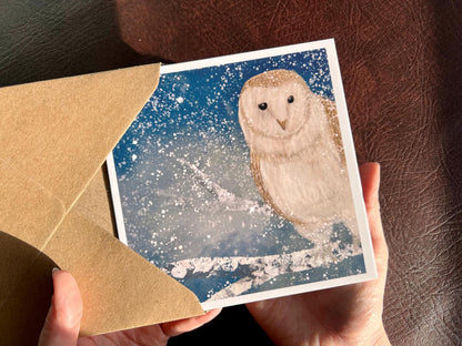 Hands holding a winter owl greeting card being removed from a recycled kraft envelope, featuring an illustrated barn owl perched on a snowy tree branch under a magical blue and green night sky with falling snow, an ethereal winter animal art greeting card.