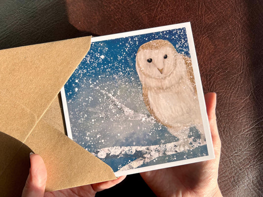 Hands holding a winter owl greeting card being removed from a recycled kraft envelope, featuring an illustrated barn owl perched on a snowy tree branch under a magical blue and green night sky with falling snow, an ethereal winter animal art greeting card.