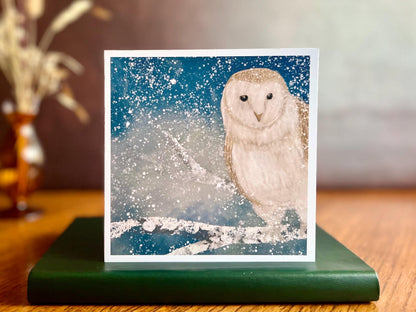 Winter owl greeting card standing on a green leather book placed on a wooden tabletop, featuring an illustrated barn owl perched on a snowy branch under a magical blue and green night sky with falling snow, an ethereal winter animal art card in a styled product photograph.