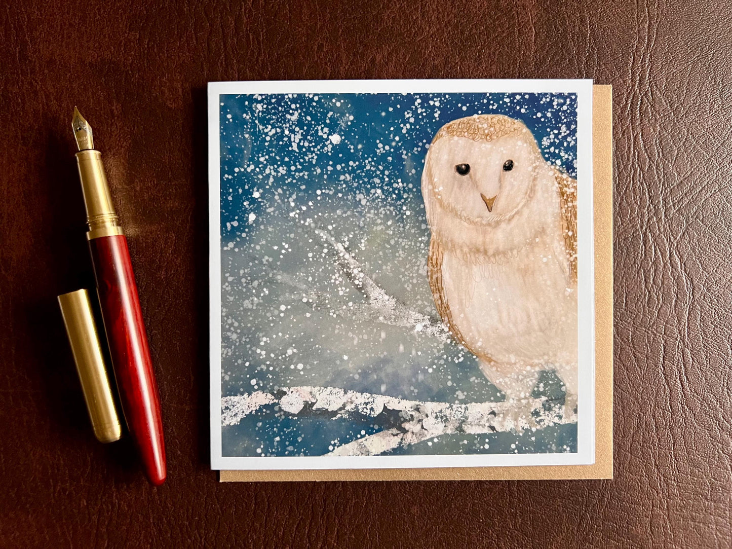 Winter owl greeting card laid flat on a leatherette tabletop with a recycled kraft envelope slotted inside the card and a fountain pen placed to the left, featuring an illustrated barn owl on a snowy branch under a magical blue and green night sky with falling snow, ethereal winter animal art greeting card.