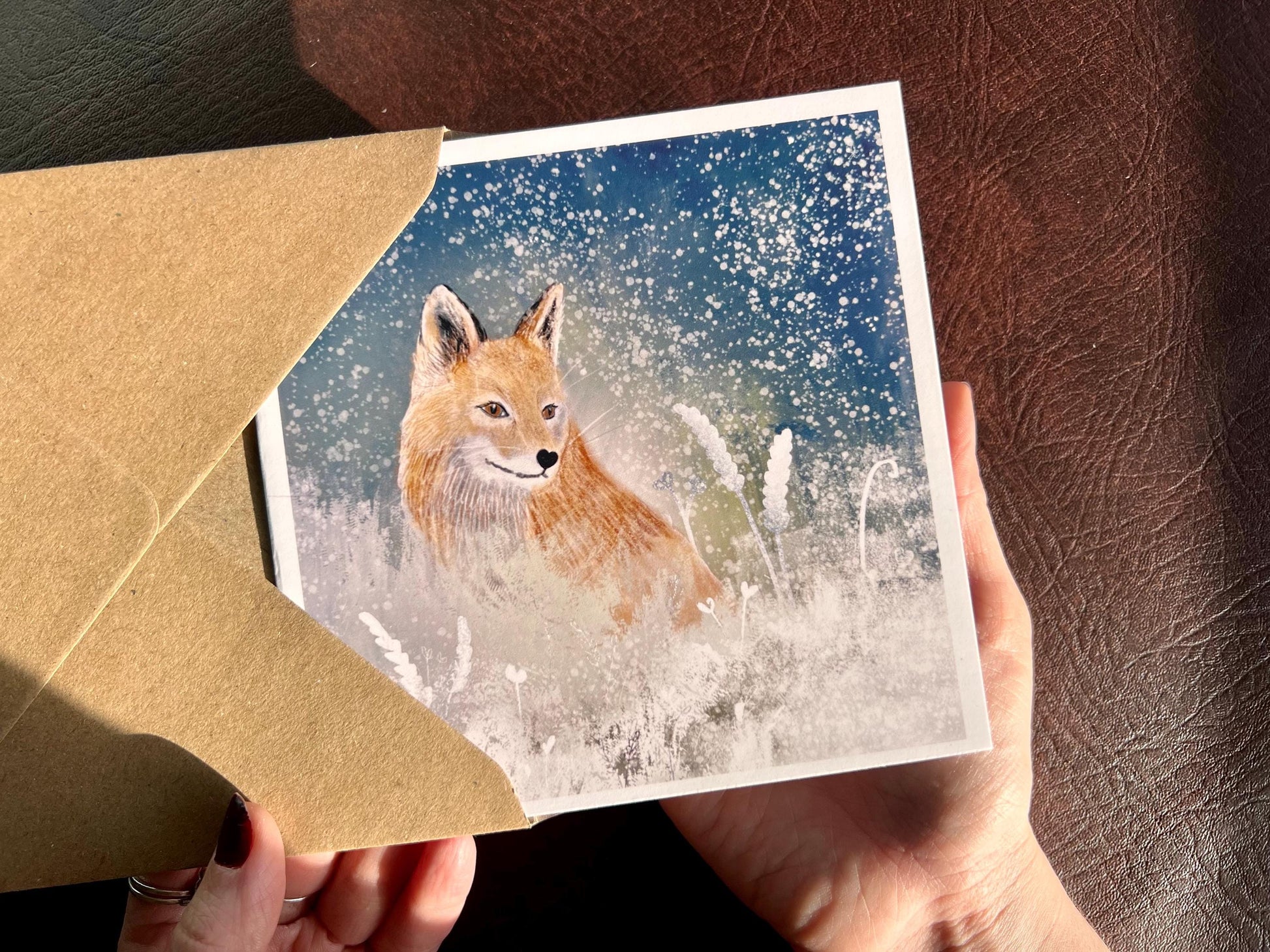 Premium winter art card featuring an illustrated woodland fox standing in a snowy night scene with falling snow, snowy foliage and a dark blue and green starry sky. The card has no text and is shown in close-up being held in two hands as it is removed from a recycled kraft envelope. Ideal seasonal greeting card for fox lovers, wildlife enthusiasts and nature inspired winter gifting. Printed on thick 312gsm fine art card by a UK independent artist.