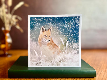 Premium winter art card featuring an illustrated woodland fox standing in a snowy night scene with falling snow, snowy foliage and a dark blue and green starry sky. The card has no text and is shown standing on a book in a styled display. Ideal seasonal greeting card for fox lovers, wildlife enthusiasts and nature inspired winter gifting. Printed on thick 312gsm fine art card by a UK independent artist.