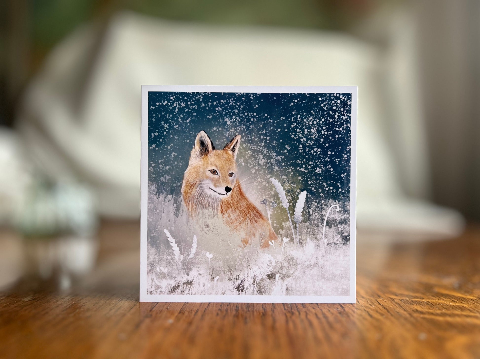 Premium winter art card featuring an illustrated woodland fox standing in a snowy night scene with falling snow, snowy foliage and a dark blue and green starry sky. The card has no text and is shown standing on a rustic wooden tabletop. Ideal seasonal greeting card for fox lovers, wildlife enthusiasts and nature inspired winter gifting. Printed on thick 312gsm fine art card by a UK independent artist.
