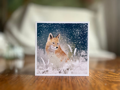 Premium winter art card featuring an illustrated woodland fox standing in a snowy night scene with falling snow, snowy foliage and a dark blue and green starry sky. The card has no text and is shown standing on a rustic wooden tabletop. Ideal seasonal greeting card for fox lovers, wildlife enthusiasts and nature inspired winter gifting. Printed on thick 312gsm fine art card by a UK independent artist.