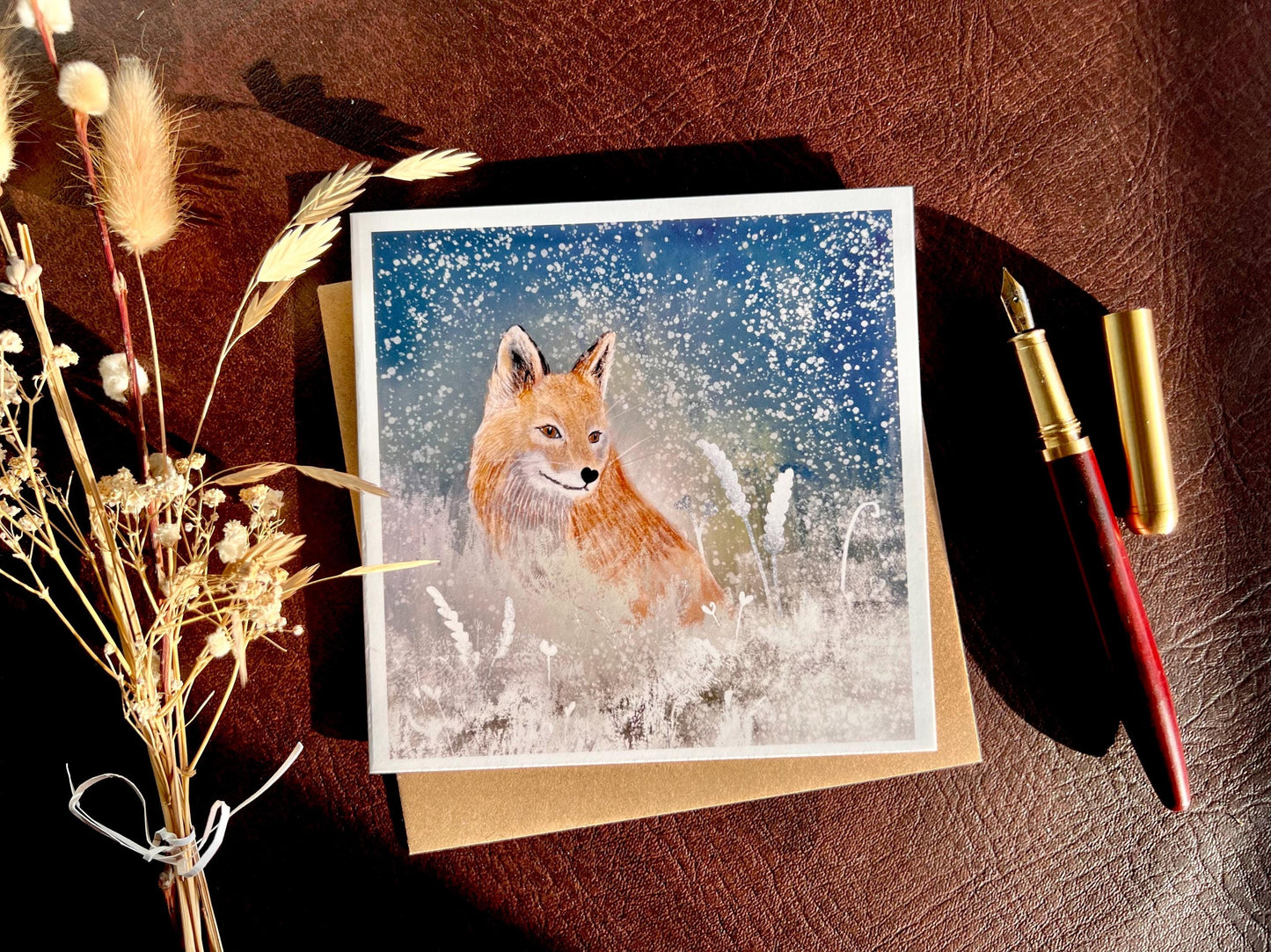 Premium winter art card featuring an illustrated woodland fox standing in a snowy night scene with falling snow, snowy foliage and a dark blue and green starry sky. The card has no text and is shown laid flat with a recycled kraft envelope and dried flowers on a tabletop, with sunlight casting soft shadows. Ideal seasonal greeting card for fox lovers, wildlife enthusiasts and nature inspired winter gifting. Printed on thick 312gsm fine art card by a UK independent artist.