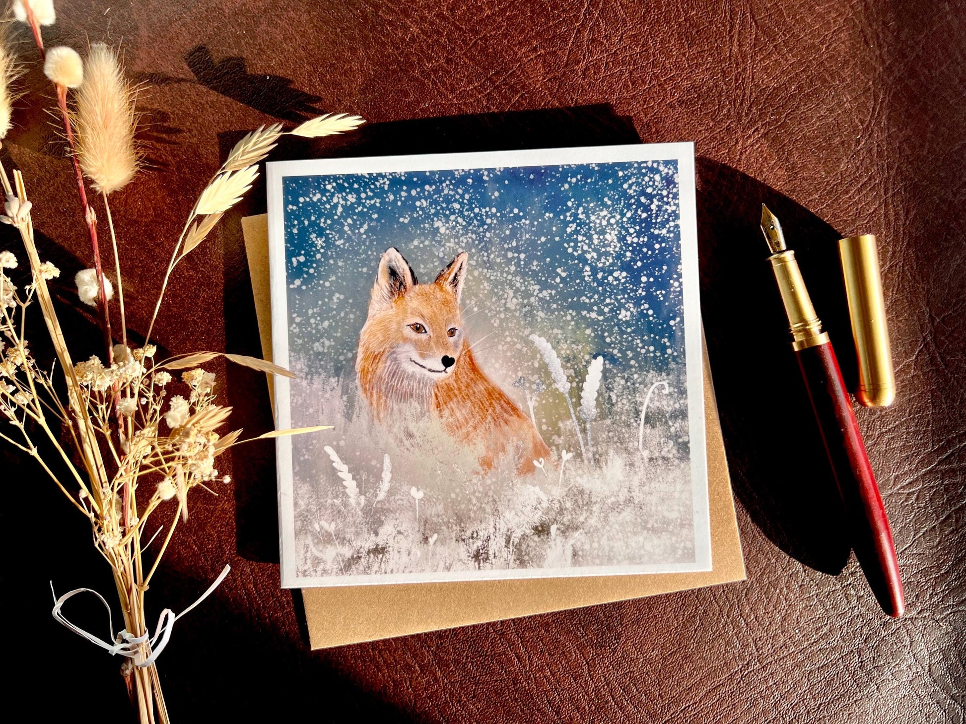 Premium winter art card featuring an illustrated woodland fox standing in a snowy night scene with falling snow, snowy foliage and a dark blue and green starry sky. The card has no text and is shown laid flat with a recycled kraft envelope and dried flowers on a tabletop, with sunlight casting soft shadows. Ideal seasonal greeting card for fox lovers, wildlife enthusiasts and nature inspired winter gifting. Printed on thick 312gsm fine art card by a UK independent artist.
