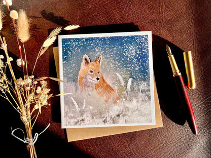 Premium winter art card featuring an illustrated woodland fox standing in a snowy night scene with falling snow, snowy foliage and a dark blue and green starry sky. The card has no text and is shown laid flat with a recycled kraft envelope and dried flowers on a tabletop, with sunlight casting soft shadows. Ideal seasonal greeting card for fox lovers, wildlife enthusiasts and nature inspired winter gifting. Printed on thick 312gsm fine art card by a UK independent artist.