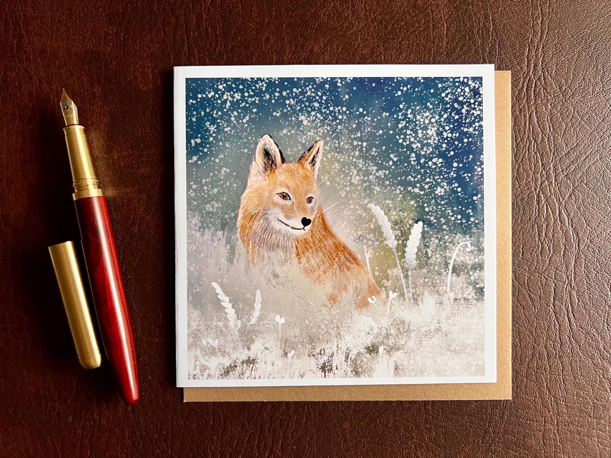 Premium winter art card featuring an illustrated woodland fox standing in a snowy night scene with falling snow, snowy foliage and a dark blue and green starry sky. The card has no text and is shown laid flat with a recycled kraft envelope and pen on a tabletop. Ideal seasonal greeting card for fox lovers, wildlife enthusiasts and nature inspired winter gifting. Printed on thick 312gsm fine art card by a UK independent artist.