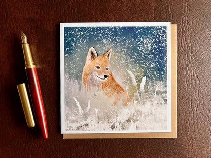 Premium winter art card featuring an illustrated woodland fox standing in a snowy night scene with falling snow, snowy foliage and a dark blue and green starry sky. The card has no text and is shown laid flat with a recycled kraft envelope and pen on a tabletop. Ideal seasonal greeting card for fox lovers, wildlife enthusiasts and nature inspired winter gifting. Printed on thick 312gsm fine art card by a UK independent artist.