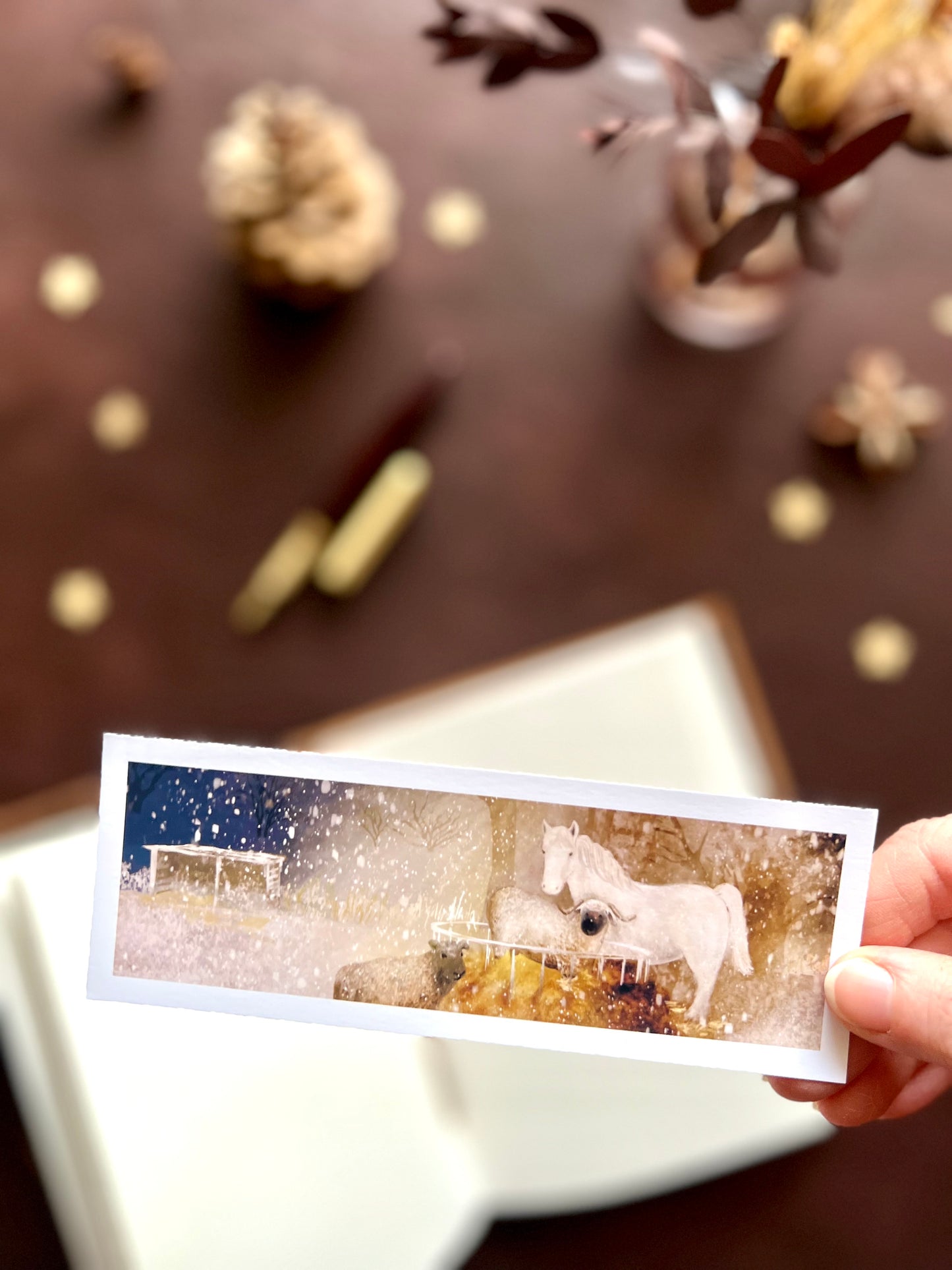 Illustrated bookmark featuring a nighttime scene with a white horse and sheep around a hay bale in the snow. Woodland themed background with sketchbook, pine cones, and kraft paper stars. Part of a 16 pack animal bookmark set for wedding favours.