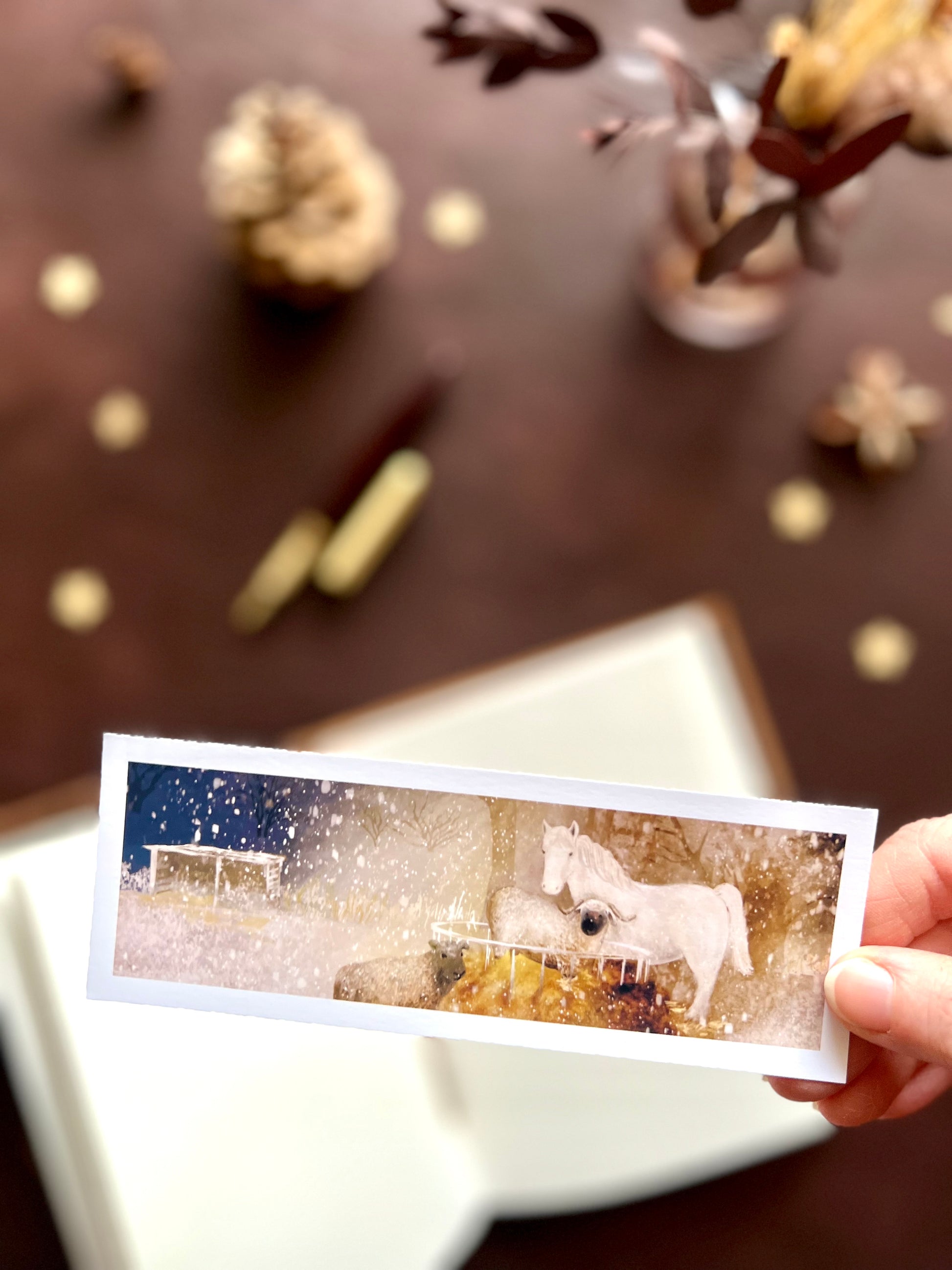 Illustrated bookmark featuring a nighttime scene with a white horse and sheep around a hay bale in the snow. Woodland themed background with sketchbook, pine cones, and kraft paper stars. Part of a 16 pack animal bookmark set for wedding favours.