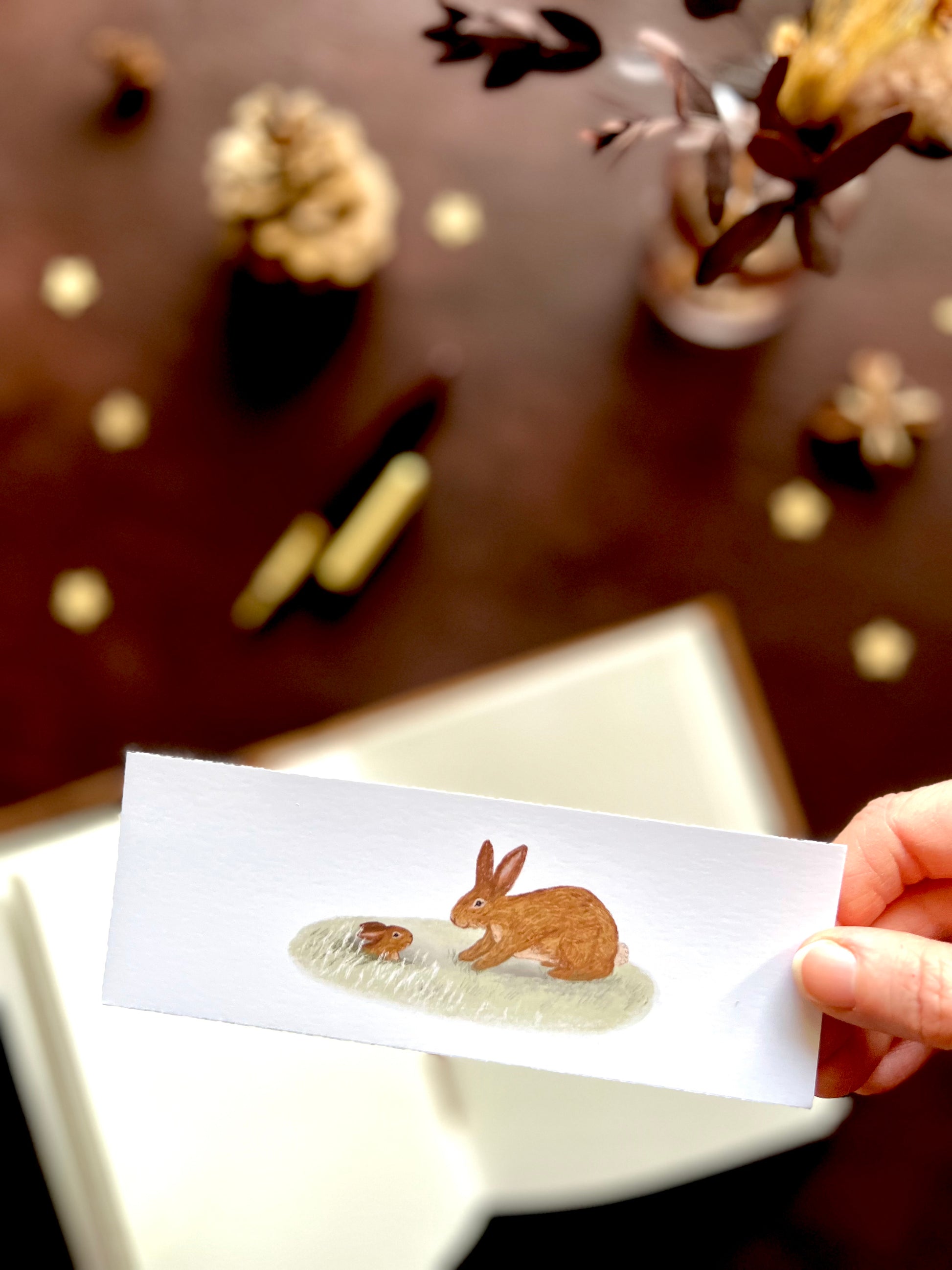 Illustrated bookmark featuring a tiny baby bunny emerging from its burrow. Woodland themed background with sketchbook, pine cones, and kraft paper stars. Part of a 16 pack animal bookmark set for wedding favours.