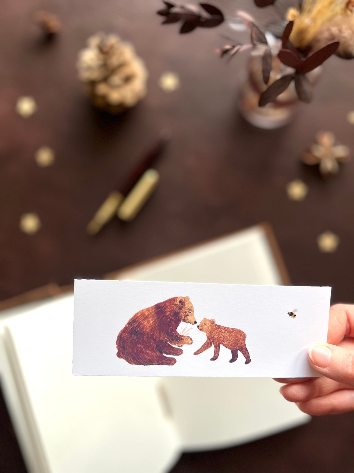 Illustrated bookmark showing a brown bear cub with its parent and a friendly bumblebee. Woodland themed background with sketchbook, pine cones, and kraft paper stars. Part of a 16 pack animal bookmark set for wedding favours.