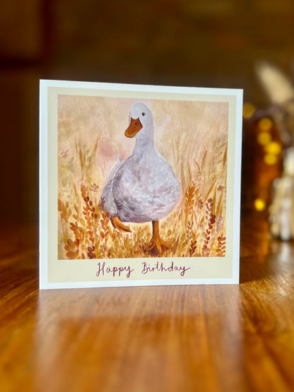 White duck Happy Birthday card standing on a rustic wooden tabletop with farmhouse decor in the background, showcasing the whimsical painted duck illustration and warm countryside feel