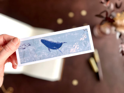 Illustrated bookmark featuring a gentle blue whale swimming through the pale blue sea. Woodland themed background with sketchbook, pine cones, and kraft paper stars. Part of a 16 pack animal bookmark set for wedding favours.