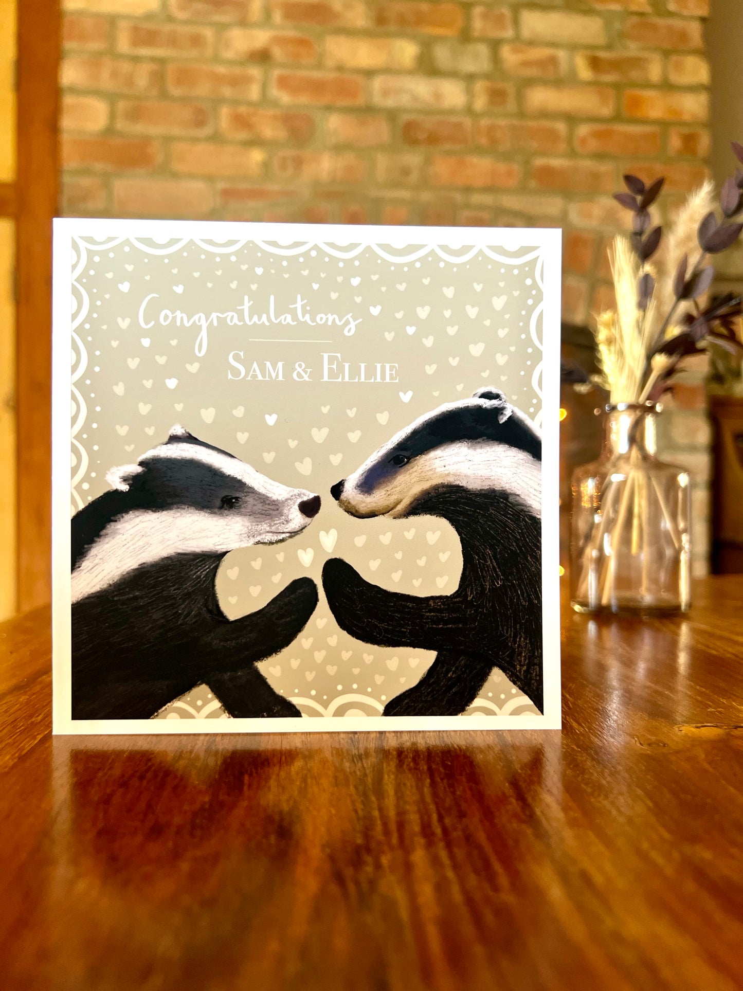 Customisable woodland wedding congratulations card with badger illustration, personalised names on sage green design, rustic wooden table with dried flowers