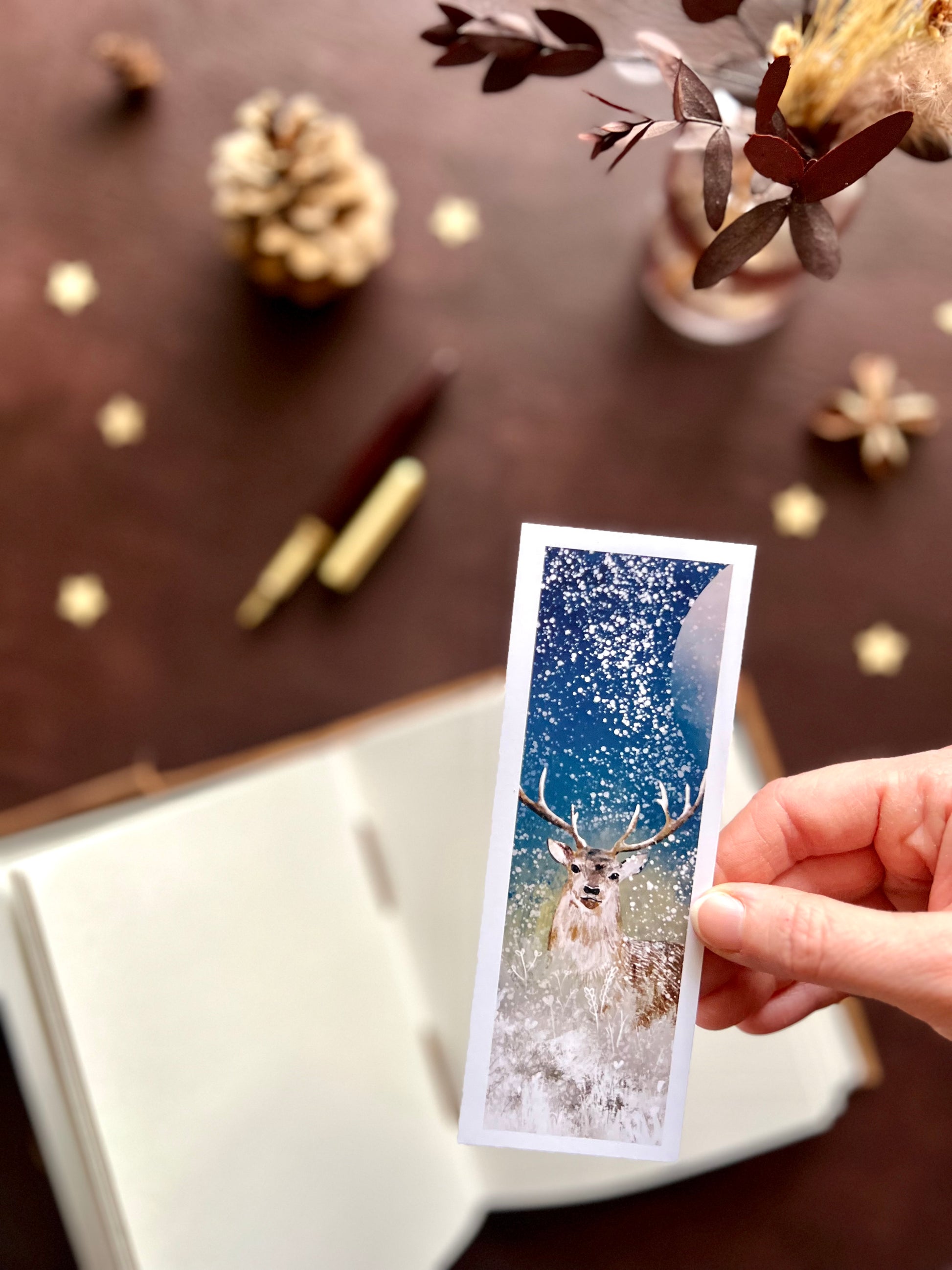 Illustrated bookmark featuring a majestic deer under the winter moon in a snowy night scene. Woodland themed background with sketchbook, pine cones, and kraft paper stars. Part of a 16 pack animal bookmark set for wedding favours.