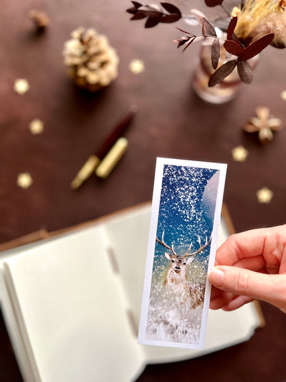 Illustrated bookmark featuring a majestic deer under the winter moon in a snowy night scene. Woodland themed background with sketchbook, pine cones, and kraft paper stars. Part of a 16 pack animal bookmark set for wedding favours.