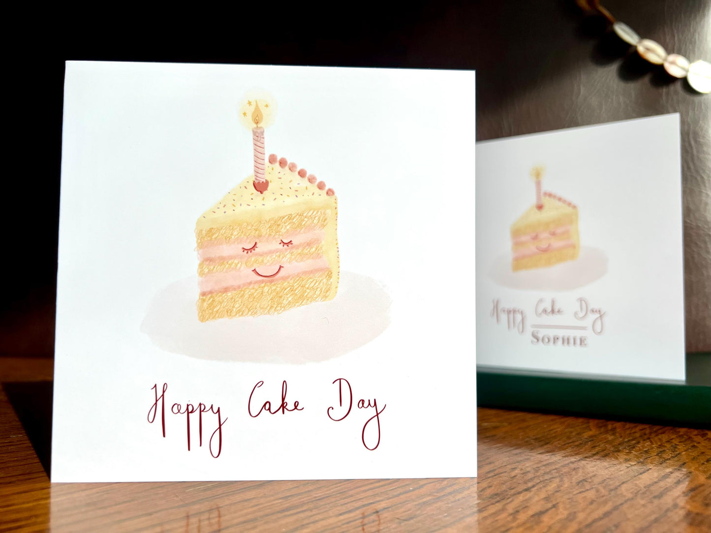 Cheerful Happy Cake Day birthday card with smiling cake illustration standing on wooden tabletop, non-personalised option shown, UK handmade