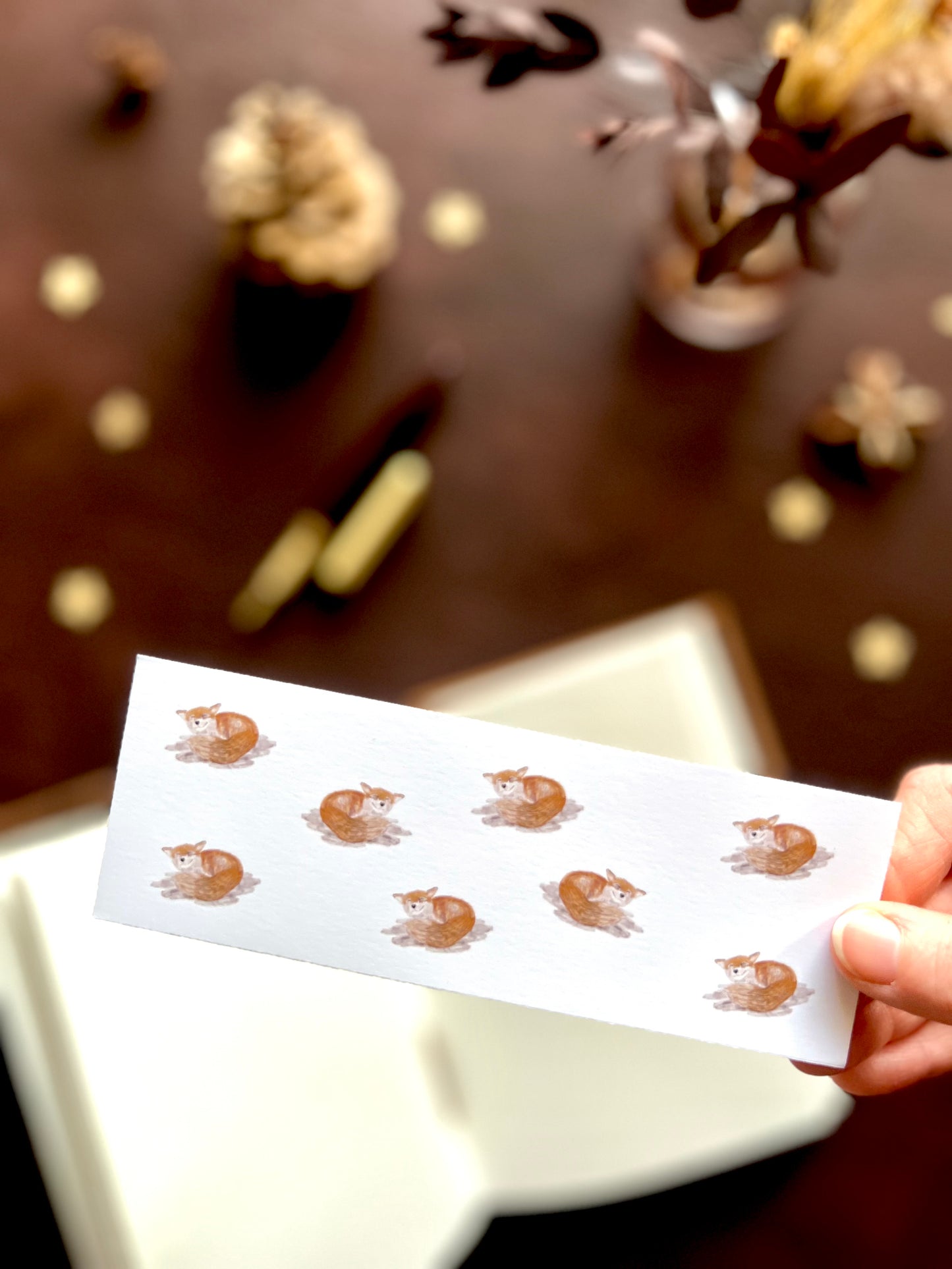 Illustrated bookmark with a repeating pattern of happy foxes held in hand. Woodland themed background with sketchbook, pine cones and kraft paper stars. Part of a 16 pack animal bookmark set for wedding favours.