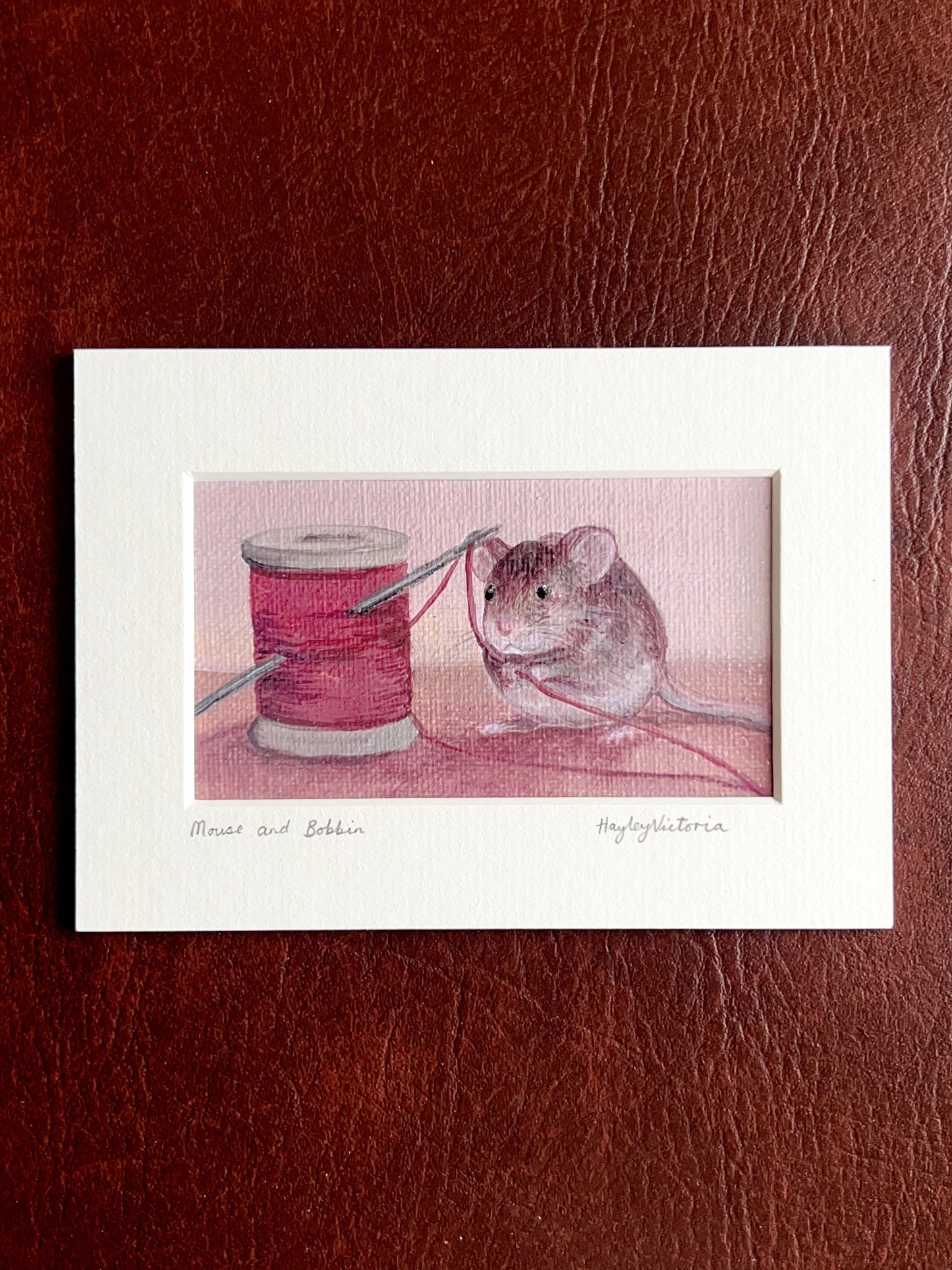 Mounted Mouse and Bobbin art print laid flat on a brown leatherette surface, showing a 5x7 landscape print with warm maroon still life painting, including handwritten title and artist signature on the mount.