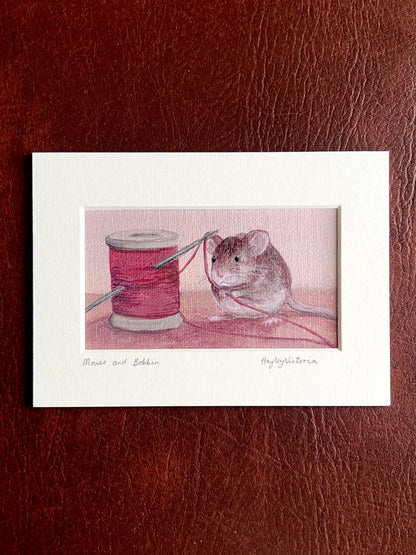 Mounted Mouse and Bobbin art print laid flat on a brown leatherette surface, showing a 5x7 landscape print with warm maroon still life painting, including handwritten title and artist signature on the mount.
