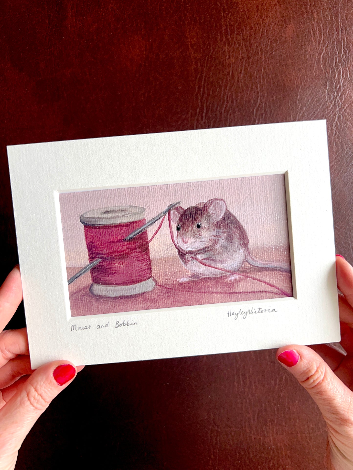 Mounted Mouse and Bobbin art print held by the artist, showing a 5x7 landscape still life painting of a mouse holding red thread beside a bobbin, demonstrating the size and detail of the artwork.