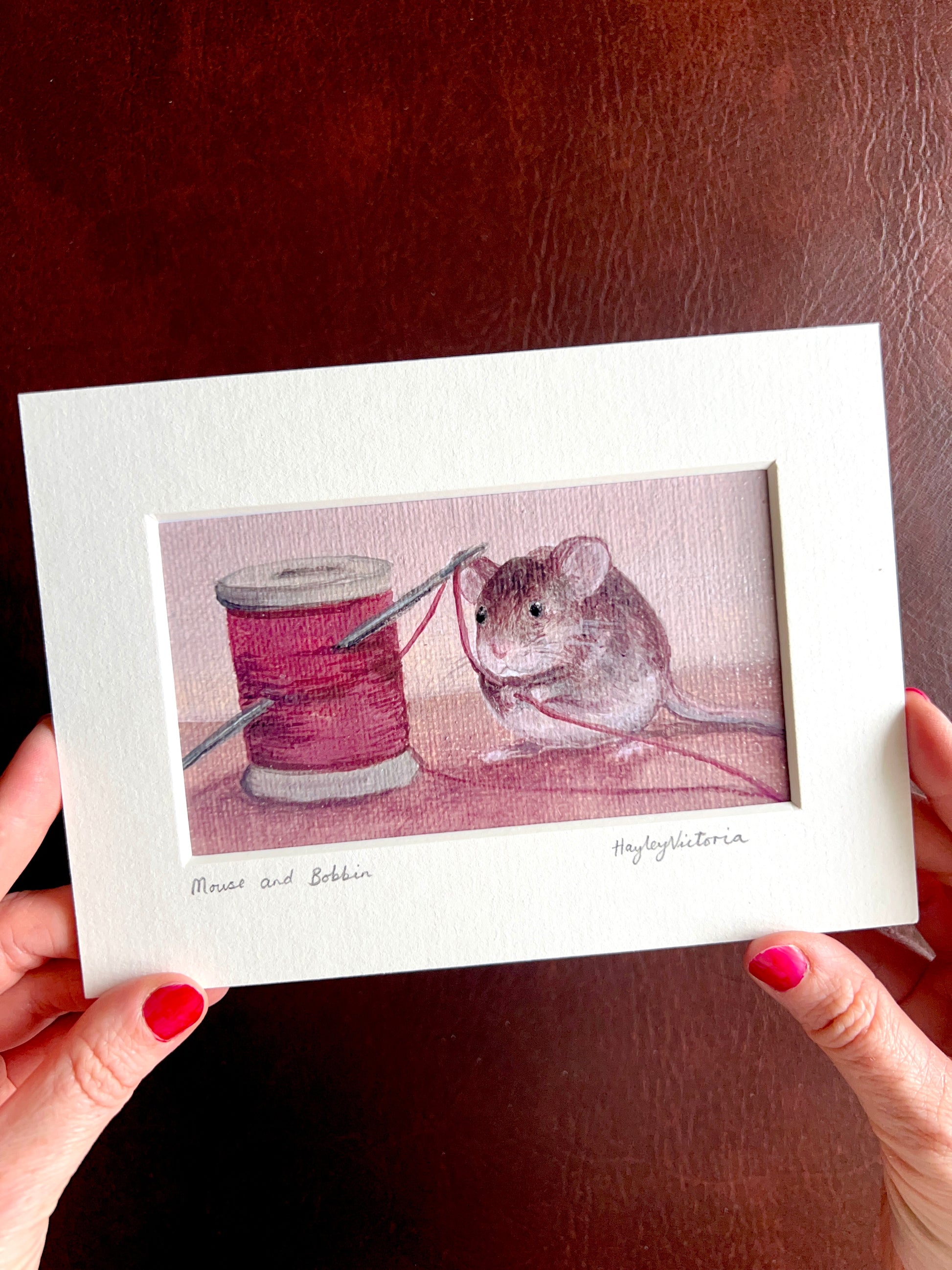 Mounted Mouse and Bobbin art print held by the artist, showing a 5x7 landscape still life painting of a mouse holding red thread beside a bobbin, demonstrating the size and detail of the artwork.