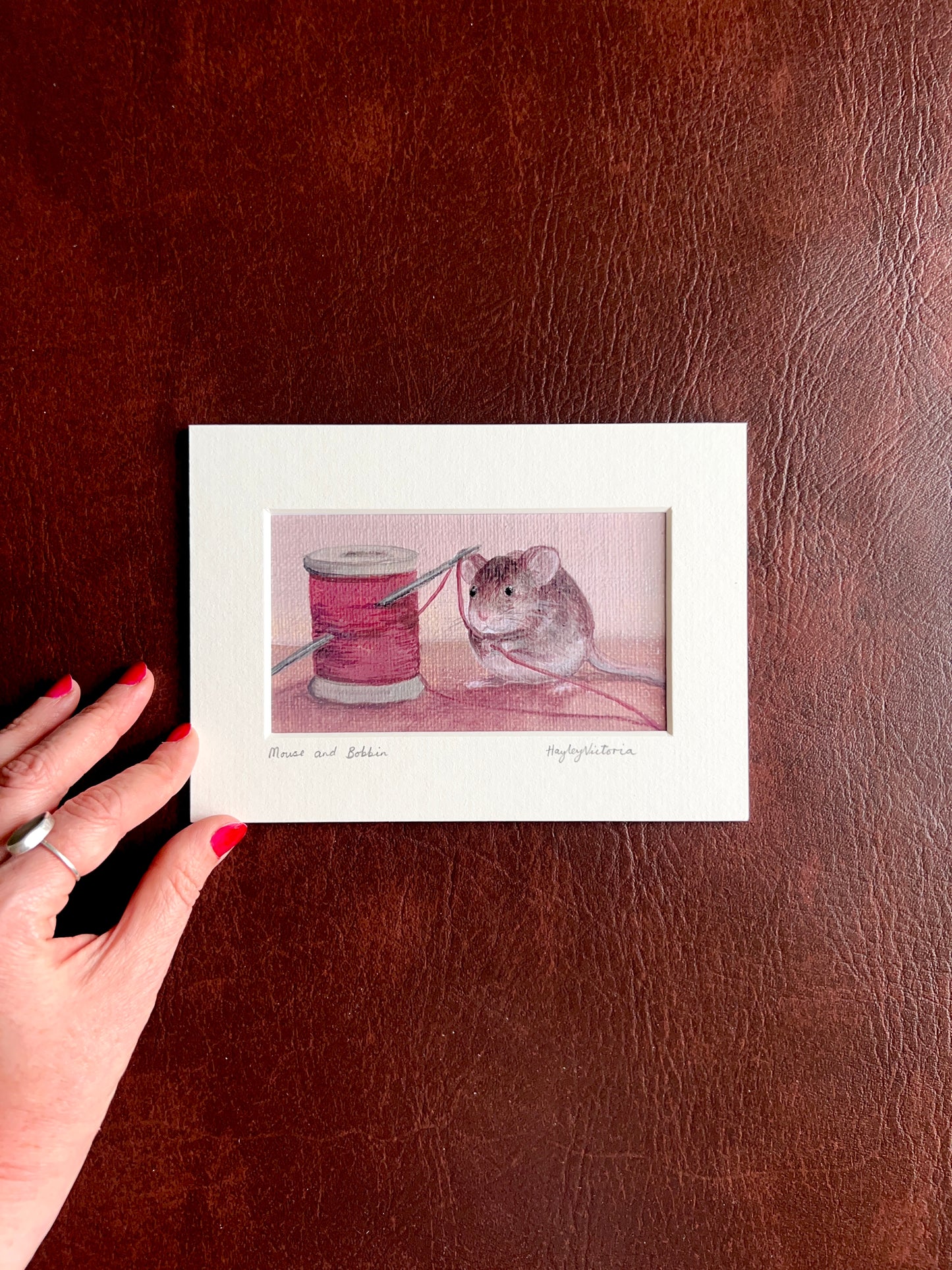 Mounted Mouse and Bobbin art print laid flat on a brown leatherette surface with a hand placed at the corner of the mount to show the size of the 5x7 landscape artwork.