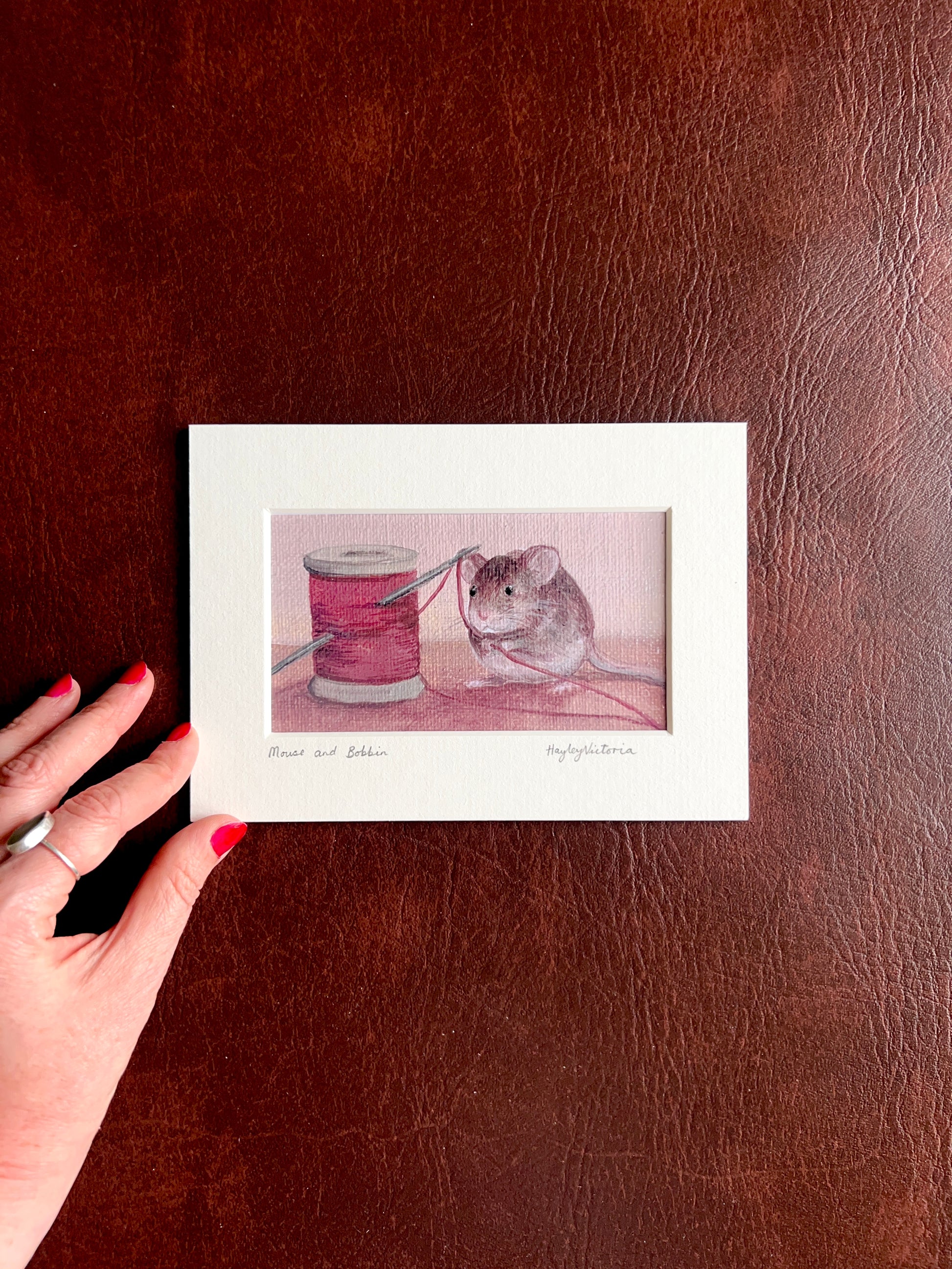 Mounted Mouse and Bobbin art print laid flat on a brown leatherette surface with a hand placed at the corner of the mount to show the size of the 5x7 landscape artwork.
