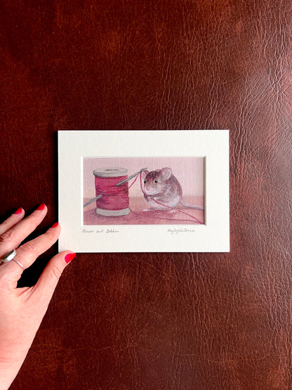 Mounted Mouse and Bobbin art print laid flat on a brown leatherette surface with a hand placed at the corner of the mount to show the size of the 5x7 landscape artwork.