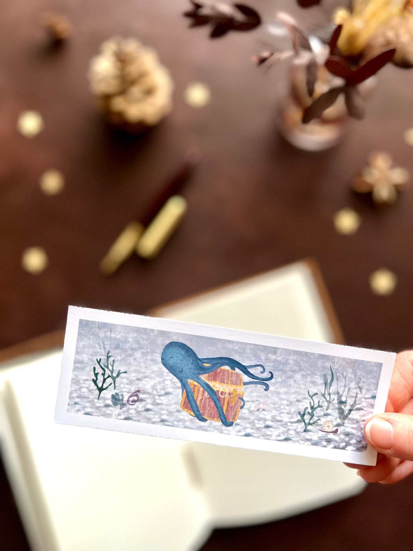 Illustrated bookmark featuring a playful blue octopus carrying treasure through the ocean. Woodland themed background with sketchbook, pine cones, and kraft paper stars. Part of a 16 pack animal bookmark set for wedding favours.