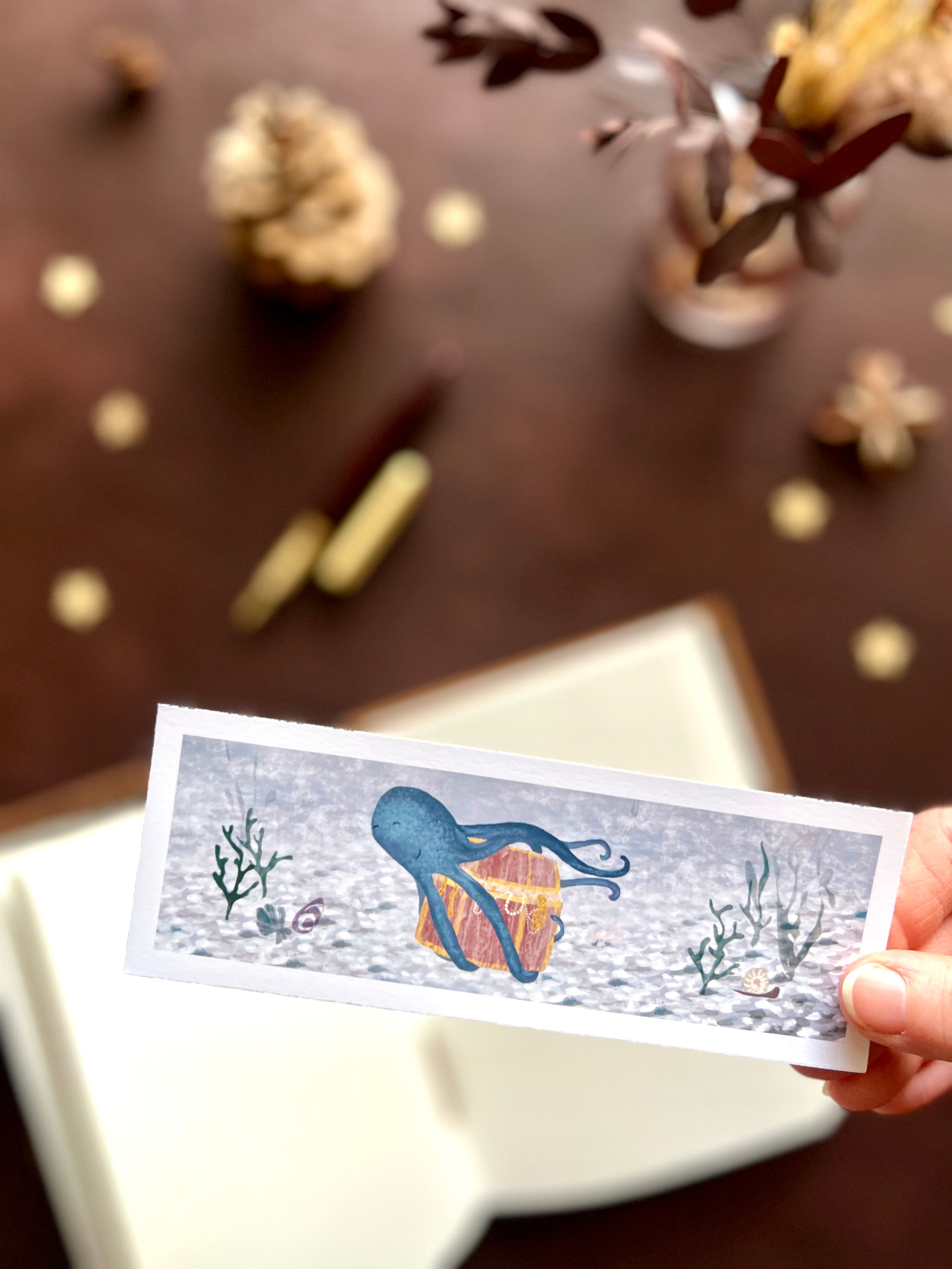 Illustrated bookmark featuring a playful blue octopus carrying treasure through the ocean. Woodland themed background with sketchbook, pine cones, and kraft paper stars. Part of a 16 pack animal bookmark set for wedding favours.