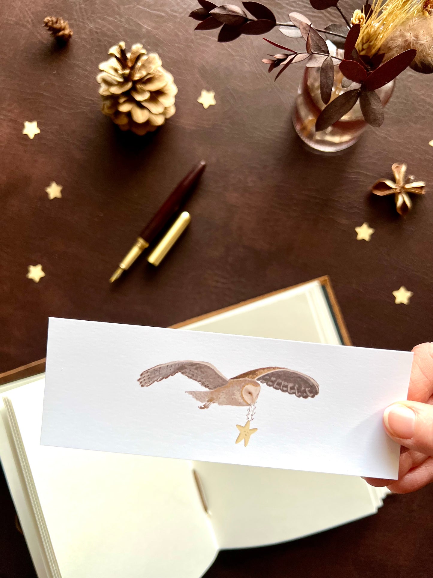 Owl illustrated bookmark with star design held in hand. Woodland themed background with sketchbook and pine cones. Part of a 16 pack animal bookmark set for wedding favours.