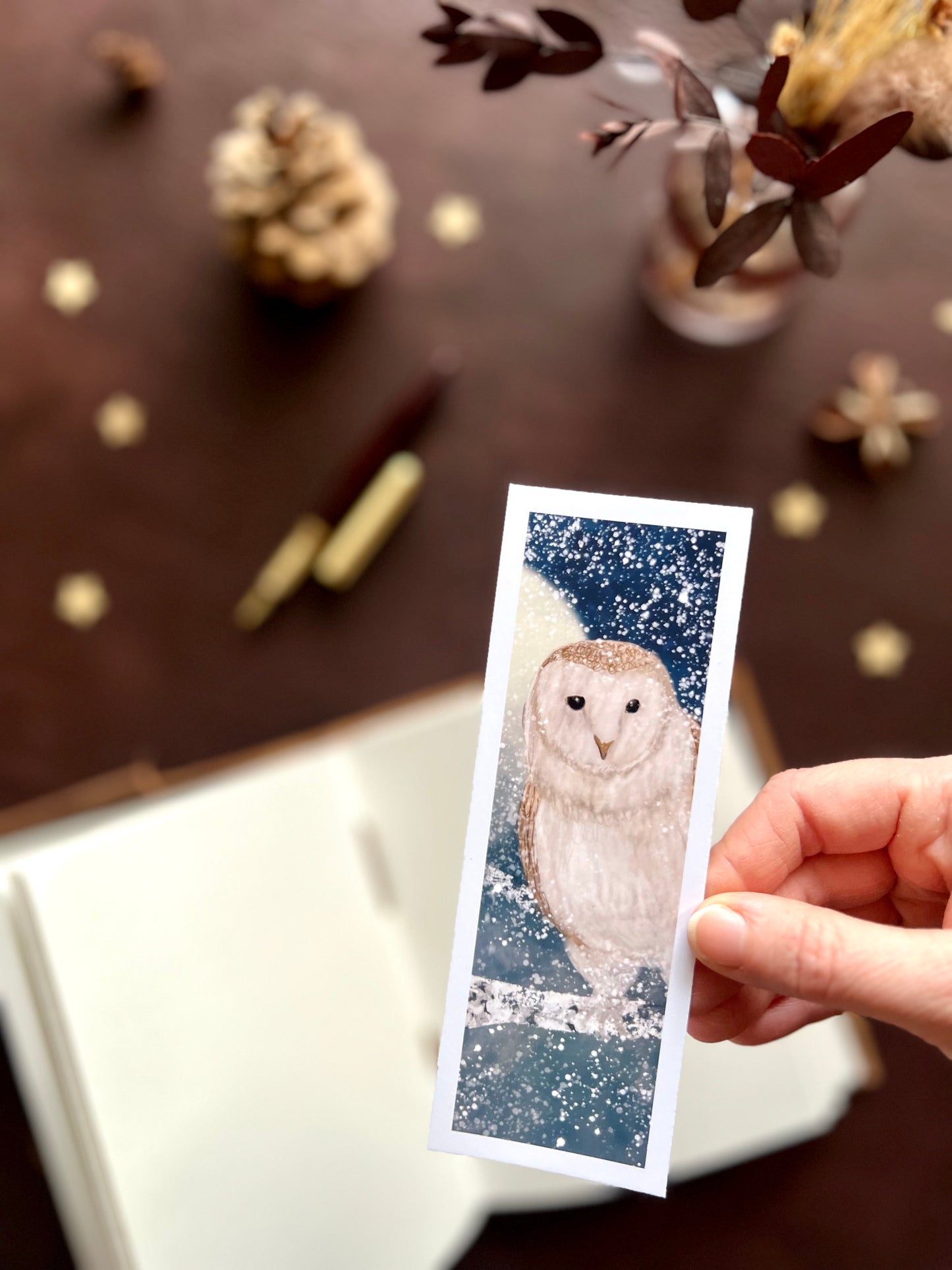 Illustrated bookmark featuring a white barn owl against a moonlit snowy night sky. Woodland themed background with sketchbook, pine cones, and kraft paper stars. Part of a 16 pack animal bookmark set for wedding favours.