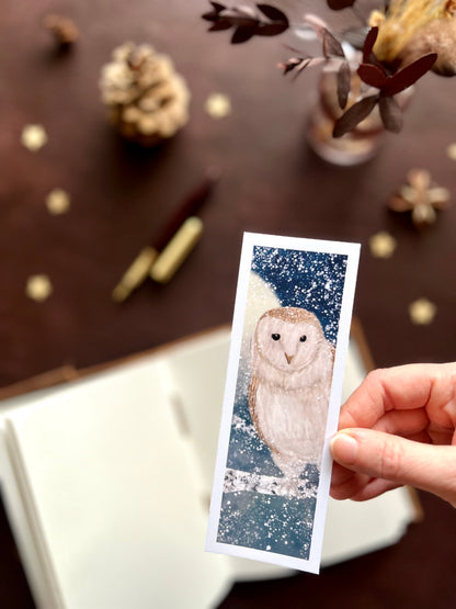 Illustrated bookmark featuring a white barn owl against a moonlit snowy night sky. Woodland themed background with sketchbook, pine cones, and kraft paper stars. Part of a 16 pack animal bookmark set for wedding favours.