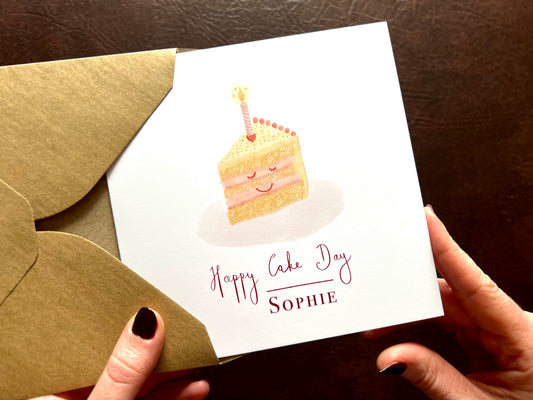 Personalised Happy Cake Day birthday card with smiling cake illustration, lemon yellow icing and pink candle, on white card being removed from 110gsm recycled kraft envelope, UK handmade