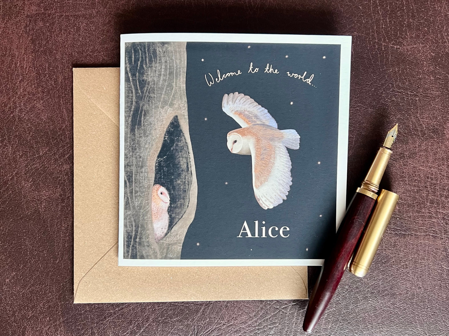 Personalised Owl New Baby Greeting Card (Premium Card) laid flat on a recycled kraft envelope, featuring a barn owl flying to its baby in a woodland night scene with handwritten text “Welcome to the World” and the personalised name at the bottom, on a matte black nighttime background, styled on a deep brown leatherette surface with a wooden fountain pen